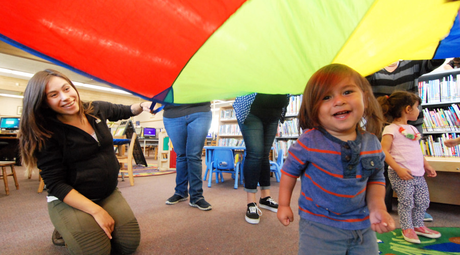 Smiling toddler under a colorful parachute as an adult looks on during playgroup.