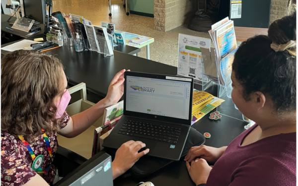 A photo of a library staff member checking out a laptop to a woman at the circulation desk at Cesar Chavez Library