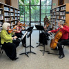 Four musicians play string instruments in a library, with bookshelves lining the walls and a large arched window in the background.