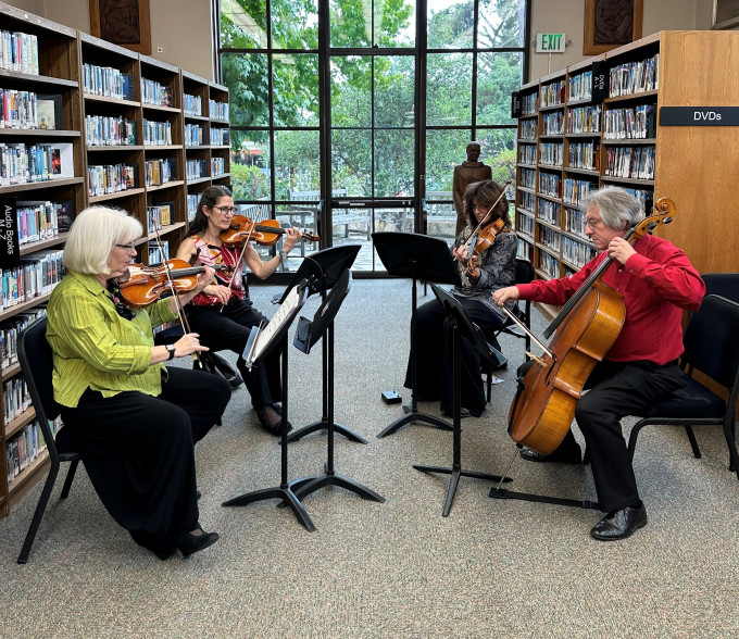 Four musicians play string instruments in a library, with bookshelves lining the walls and a large arched window in the background.