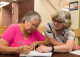 A woman writes in a workbook while a tutor leans in to help, in the Literacy Center.